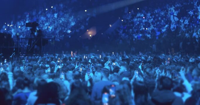 Large group of people standing, watching music concert. Spectators of show shine phones in concert hall darkness, indoor stadium. Auditorium of thousands filled by people crowd. performance event