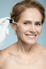 Naklejka premium Facial skin care with treatment cosmetic oil and serum. Beautiful Caucasian woman applying serum on her face, isolated on blue background