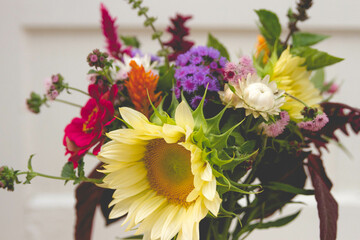 A close-up front view of a light yellow sunflower in a bouquet with strawflower, basil, zinnia, celosia and amaranth with a white door background