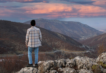 A man stands on a rock overlooking a valley and looks out at the sunset.
