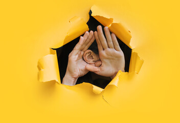 Close-up of a woman's ear and two hand through a torn hole in the paper. Yellow background, copy space. The concept of eavesdropping, espionage, gossip and tabloids. - obrazy, fototapety, plakaty