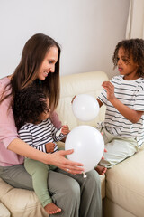 A mother shares a cheerful moment with her two African American daughters, playing with balloons on a sofa, radiating familial warmth, and mixed race families .