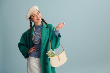 Fashionable happy smiling woman wearing beret, green coat, turtleneck, pearl necklace, holding trendy white leather bag with snowdrops, posing on blue background. Copy, empty, blank space for text - 752534125
