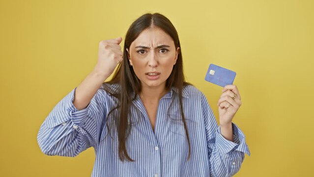 Young, beautiful hispanic woman, seething with rage, shouts in frustrated anger. she clutches a credit card, loosing a furious yell, isolated on a striking yellow background