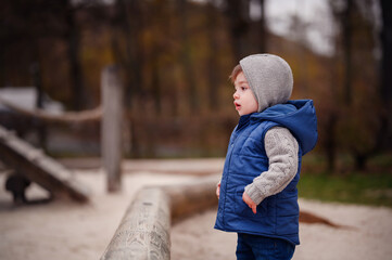A little boy in a cozy vest and knit hat experiences the joys of a playground, his expression...