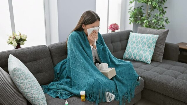 A sick woman covers her face with a tissue while sitting on a couch under a blanket with medication nearby, depicting illness at home.