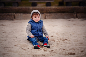 A young child dressed warmly in a vest and hat sits thoughtfully in the sand of a playground,...
