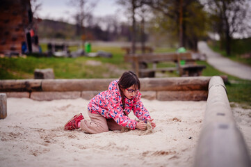 A young girl, focused and playing in the sand, sits in a playground with a beautiful, soft-focus...