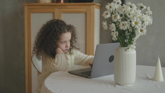 Sweet cute curly hair Bored School girl child using laptop online educational lesson course at home. Distance learning course remote video conference. Schoolgirl study does school homework in kitchen