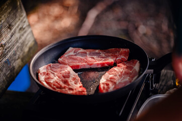 The enticing aroma of steak fills the air as it cooks in a pan on a camp stove, with wisps of smoke rising, highlighting a perfect camping meal