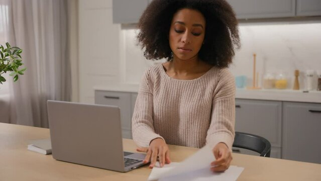 African American woman freelancer businesswoman working with papers documents at table in kitchen ethnic serious girl pay online utility bills domestic budget internet services on laptop pc at home