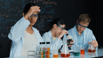 Cute girl looking under microscope while student doing experiment at blackboard with theory...