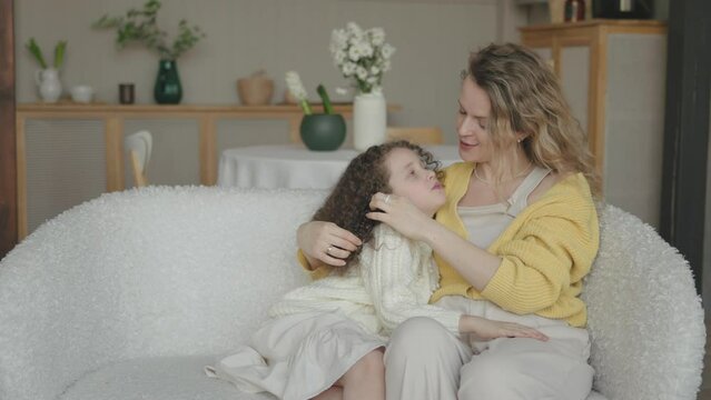 Happy little curly Cute Girl hugging, embracing her mom enjoying, bonding, and laughing together. Mother parent hugging her kid having a tender and lovely moment in living room at home