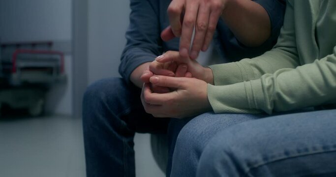 Relatives are waiting for the patient sitting on the couch in the hospital corridor and holding hands. Close-up of their hands, unrecognizable persons