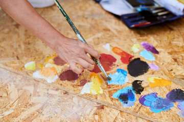Female painter hand dips paintbrush into palette of colorful paints for live painting of picture for outdoor street exhibition, close up view of female artist hand holding paintbrush