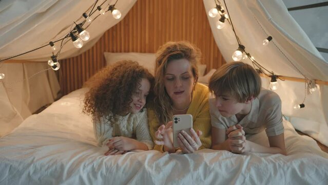 Happy caucasian mother and siblings son and daughter lying in makeshift tent with lights at night, using smartphone for doing shopping online. Communication gadget technology. Single mum and children 