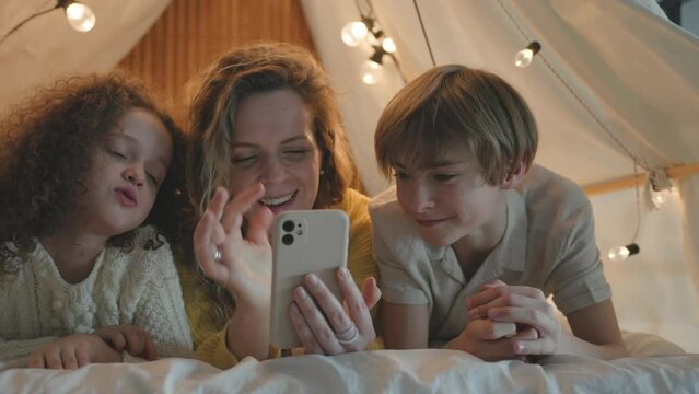 Pretty European mother spending Time with children son and daughter, doing shopping online, using smartphone, mobile phone, lying in a cozy tent house with lights in the evening. Family time with kids