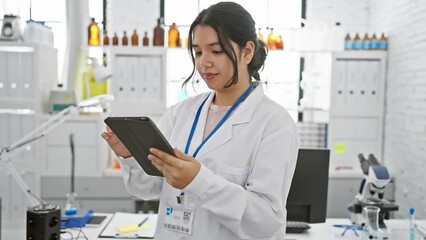 Hispanic woman scientist using tablet in laboratory setting, analyzing data amidst equipment.