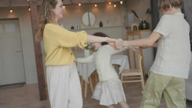 Young happy mother caucasian mommy babysitter woman holding in arms hands little daughter and son, child kid small girl boy dancing together in living room to music whirl spinning sing song having fun