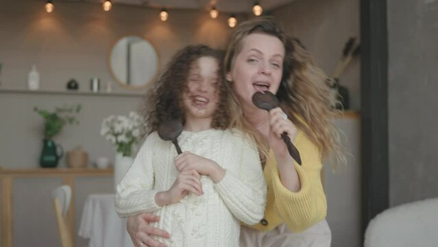 Mother and her little curly daughter having fun singing karaoke with spoons in the kitchen. Happy smiling mum enjoy family leisure time with little girl at home. Concept of having fun together.