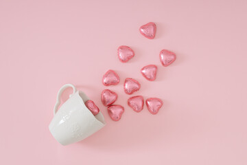 Creative love composition made with pink foil wrapped chocolate hearts coming out of white coffee cup on pastel pink background. Minimal love concept. Romantic chocolate hearts idea.