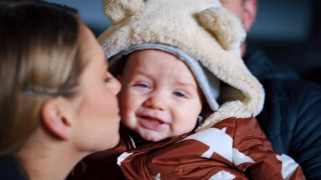 Man holding a little kid wearing warm brown and white jacket. Mother wants to kiss a baby and he gets nervous.