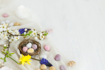 Colorful easter chocolate eggs in nest, spring flowers, feathers and linen border composition on white wooden table. Space for text. Happy Easter! Seasons greetings