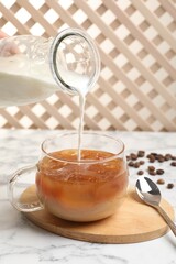Woman pouring milk into glass with refreshing iced coffee at white marble table, closeup