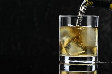 Pouring tasty whiskey from bottle into glass at mirror table against black background, closeup. Space for text