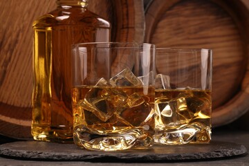 Whiskey with ice cubes in glasses, bottle and wooden barrels on black table