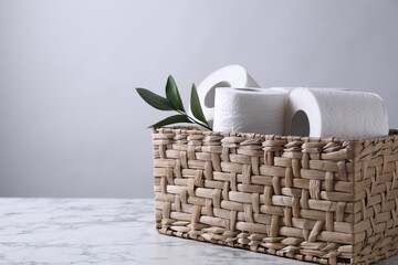 Toilet paper rolls and green leaves in wicker basket on white marble table, space for text