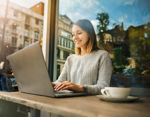 Smiling young woman working on her laptop computer in a cafe with large windows. - 752583527