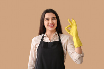 Young woman in apron and rubber gloves showing OK on beige background