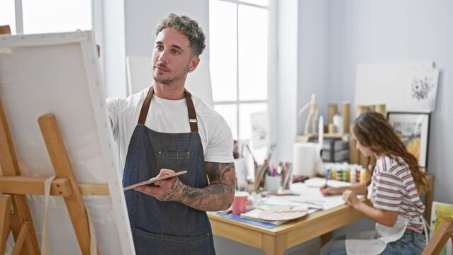 A tattooed man and a woman work in a bright art studio, focused on painting and sketching.