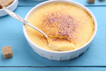 Delicious creme brulee in bowl, sugar cube and spoon on light blue wooden table, closeup