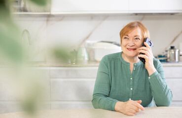 Fototapeta premium Adult positive woman in casual clothes talking on mobile phone in kitchen
