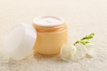 Fototapeta na wymiar Body care. Moisturizing cream in open jar and flower on light textured table, closeup