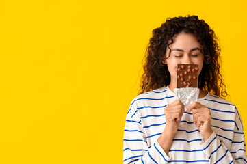 Beautiful young African-American woman with sweet chocolate bar on yellow background