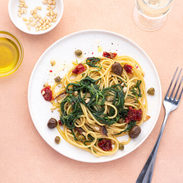 Spaghetti with Italian Agretti, Barba di frate or Saltwort or Salsola Soda, olives, anchovy, tomatoes, capers,&nbsp;&nbsp;pine nuts and olive oil, spring Italian recipes. Pink soft background, square crop