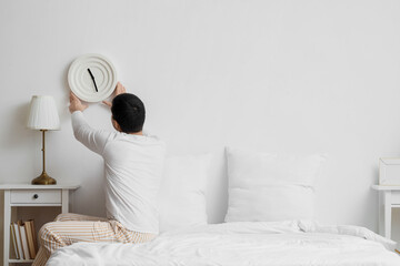 Young man with clock on light wall in bedroom, back view
