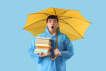 Shocked young man in raincoat with umbrella and books on blue background