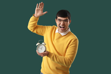 Angry young man with alarm clock on green background
