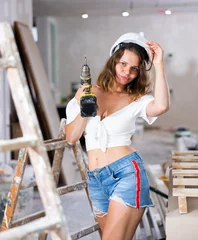 Foto op Canvas Sexy woman in a white blouse and short shorts poses playfully with a screwdriver in a renovated room © JackF