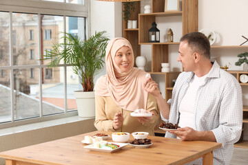 Happy Muslim woman with her husband sitting at table with traditional sweets in living room. Eid al-Fitr celebration