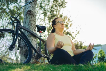 Tuinposter middle-aged latin woman sitting in a park in lotus position meditating next to her bicycle, mindfulness. copy space © juanpablo