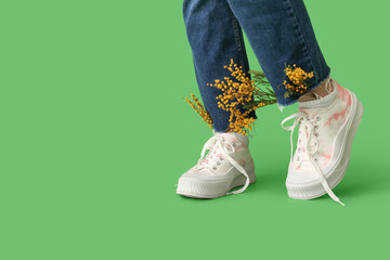 Legs of beautiful young woman in sneakers with mimosa flowers on green background
