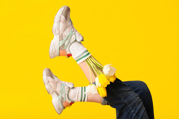 Legs of beautiful young woman in sneakers with tulips flowers on yellow background