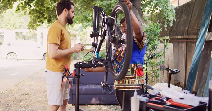 Committed interracial couple outside servicing bicycle components. Healthy active black woman and caucasian man inspecting and maintaining bike tires and chains for leisure cycling. Pull-back shot.