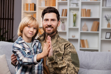 Father in army uniform, giving a high five to his small son. They're sitting and hugging on couch