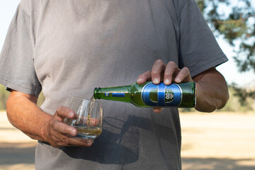 Man pouring a bottle of cold blue label  0.0 alcohol free beer into a glass on a hot sunny day, sun shadow on t-shirt